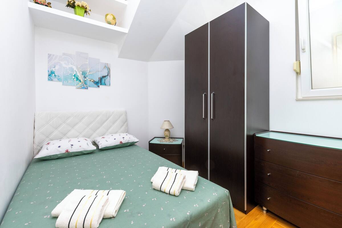 A bedroom with a bed, pillows, a nightstand, a dresser, and artwork on the wall.