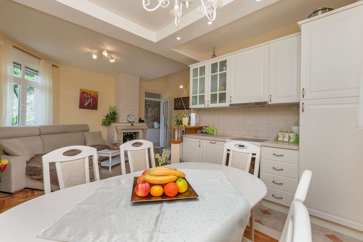 A modern kitchen with white cabinets, a dining table with a fruit centerpiece, and a living area with a sofa and chairs.