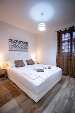 A neatly made bed with pillows and towels, a bedside table with a lamp, a wall-mounted artwork, and a window with curtains. - Apartments Ivana (Supetarska Draga)