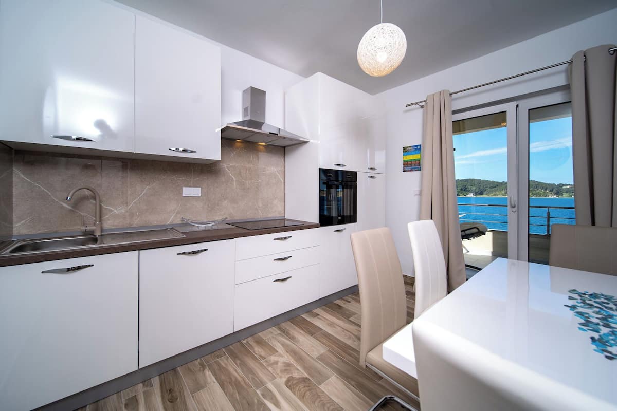 Une cuisine moderne avec des armoires blanches, un évier en acier inoxydable et un four encastré. On y trouve un coin repas avec une table et des chaises blanches, et une vue sur la mer par une grande fenêtre.