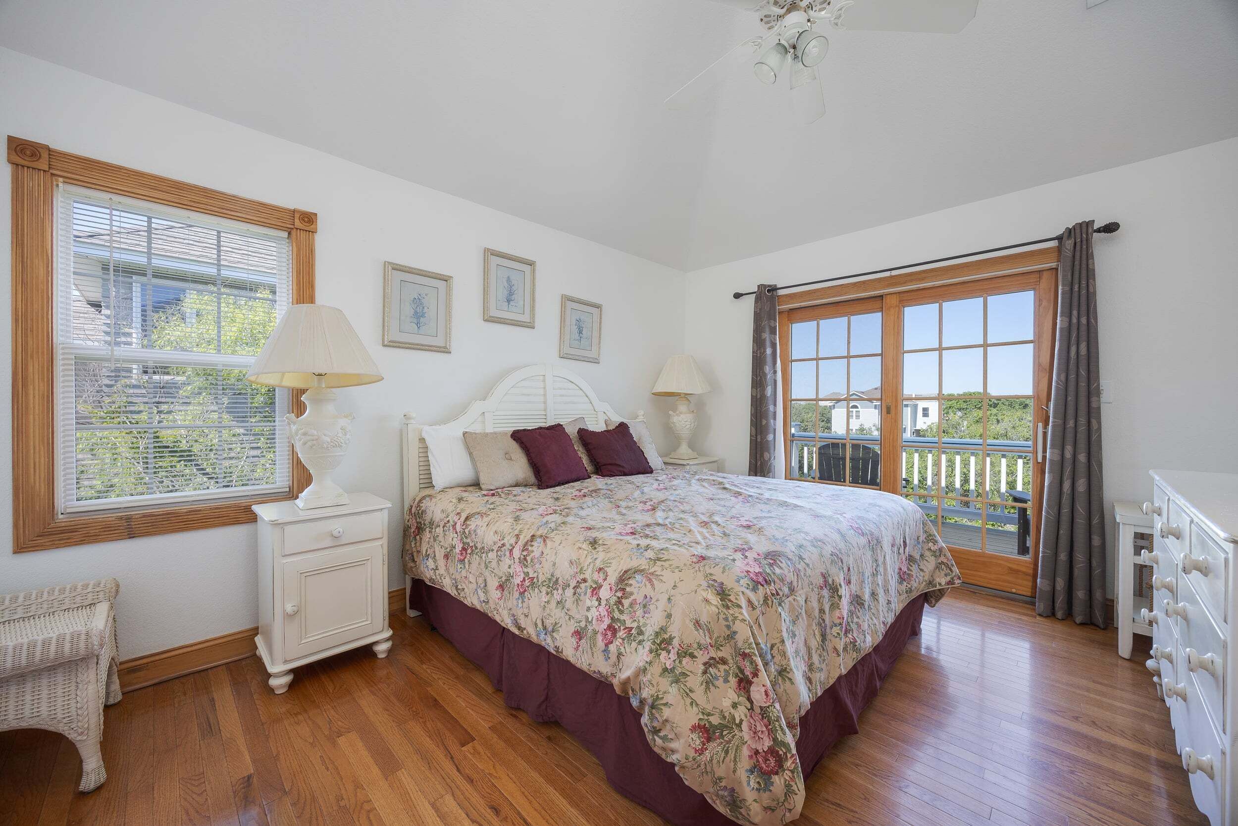 A bedroom with a bed, two bedside tables, a nightstand, a window with blinds, a balcony door, and a view of the outside.