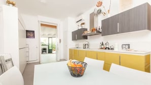 Une cuisine moderne avec des meubles blancs et jaunes, une table à manger blanche et un panier de fruits posé sur la table.
