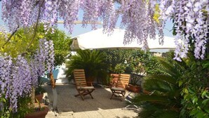 Un espace patio avec des chaises en bois et un parasol blanc, entouré d’une végétation luxuriante et de fleurs violettes.