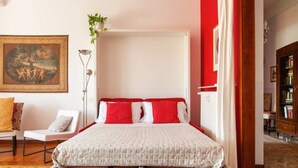 A bedroom with a white bed, red and white pillows, a painting on the wall, a chair, and a bookshelf. - Dream House (Milano)