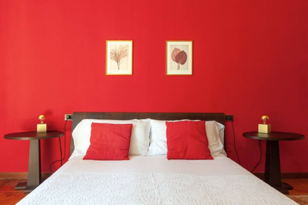 A hotel room with a red wall, a bed with white and red bedding, and two framed pictures on the wall.