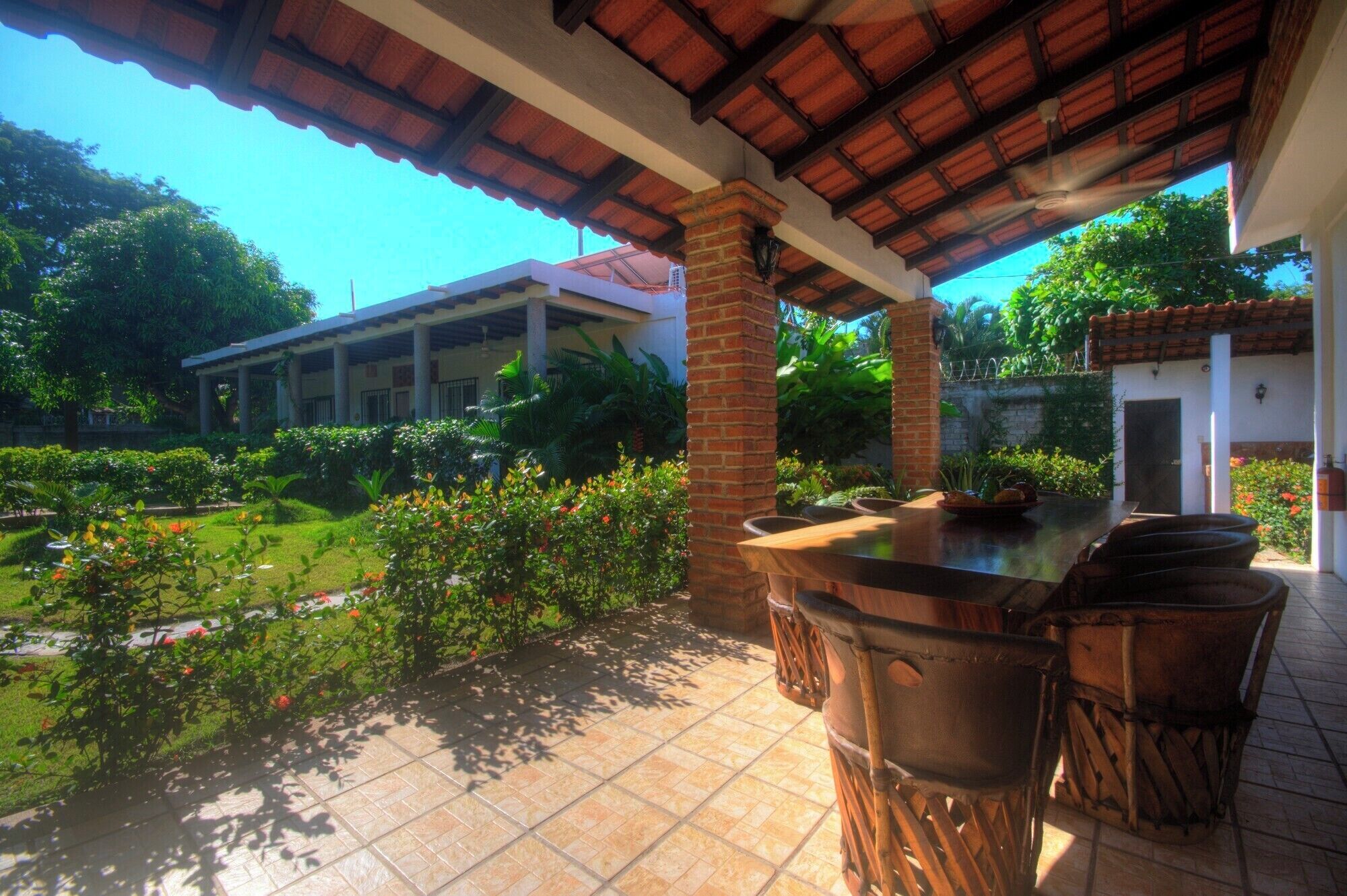 A covered patio with a dining table and chairs, surrounded by lush greenery and a view of a building in the background.