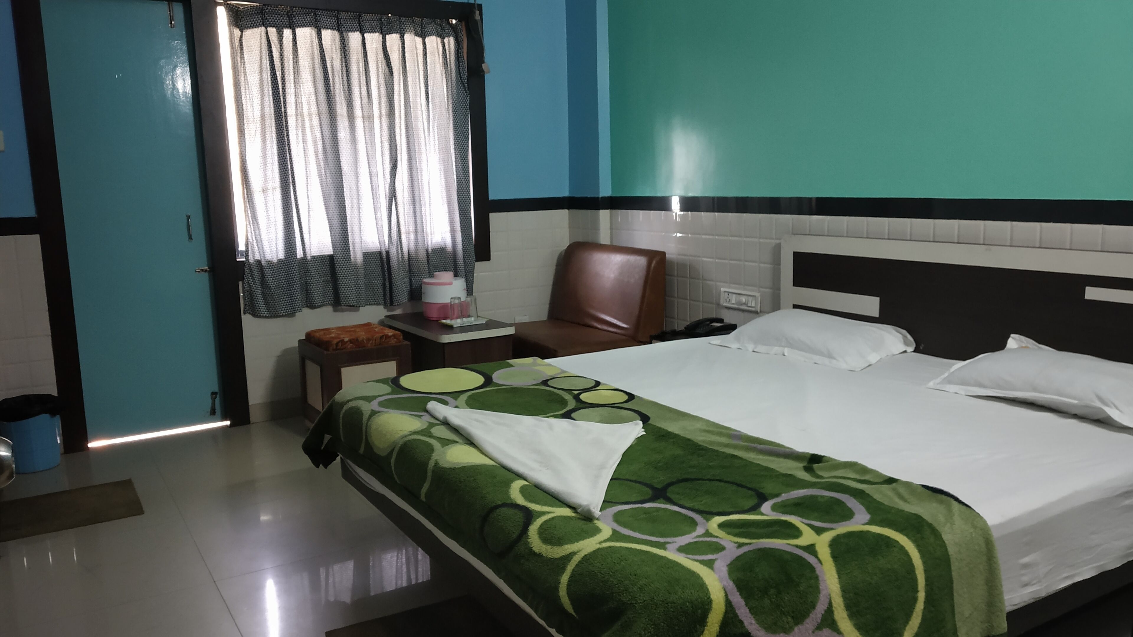 A hotel room with a bed, a chair, a small table, a nightstand, a window with curtains, and a wall with a teal and white color scheme.