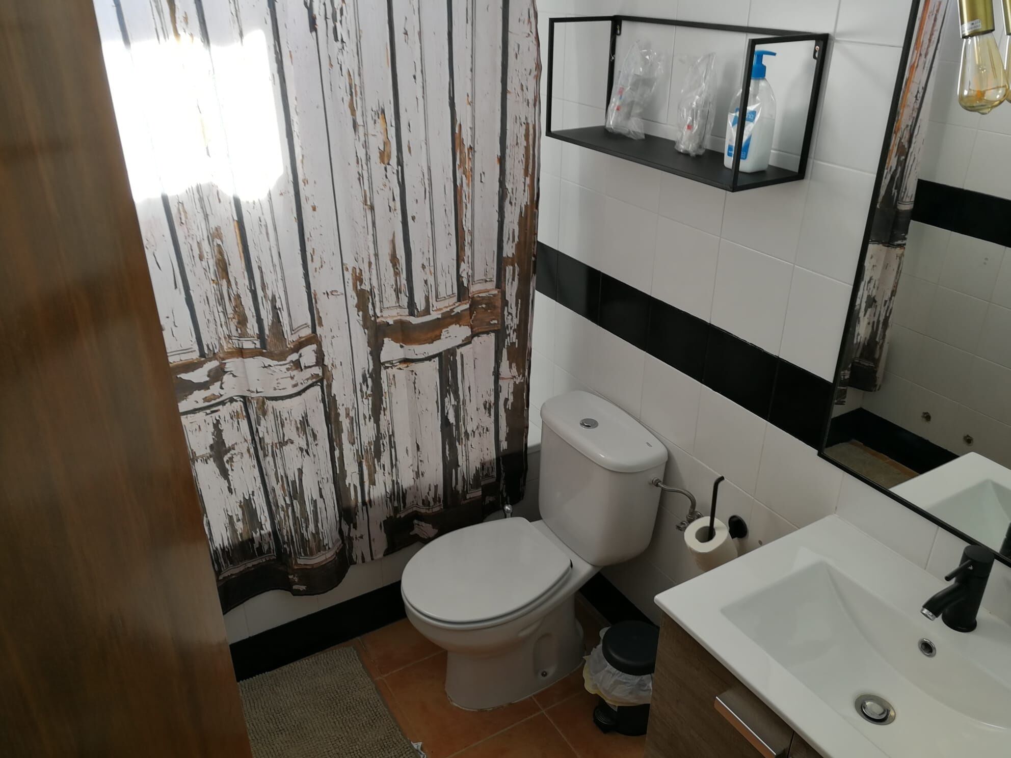 Un baño con inodoro blanco, lavamanos y espejo. Hay un estante sobre el inodoro con artículos de aseo.