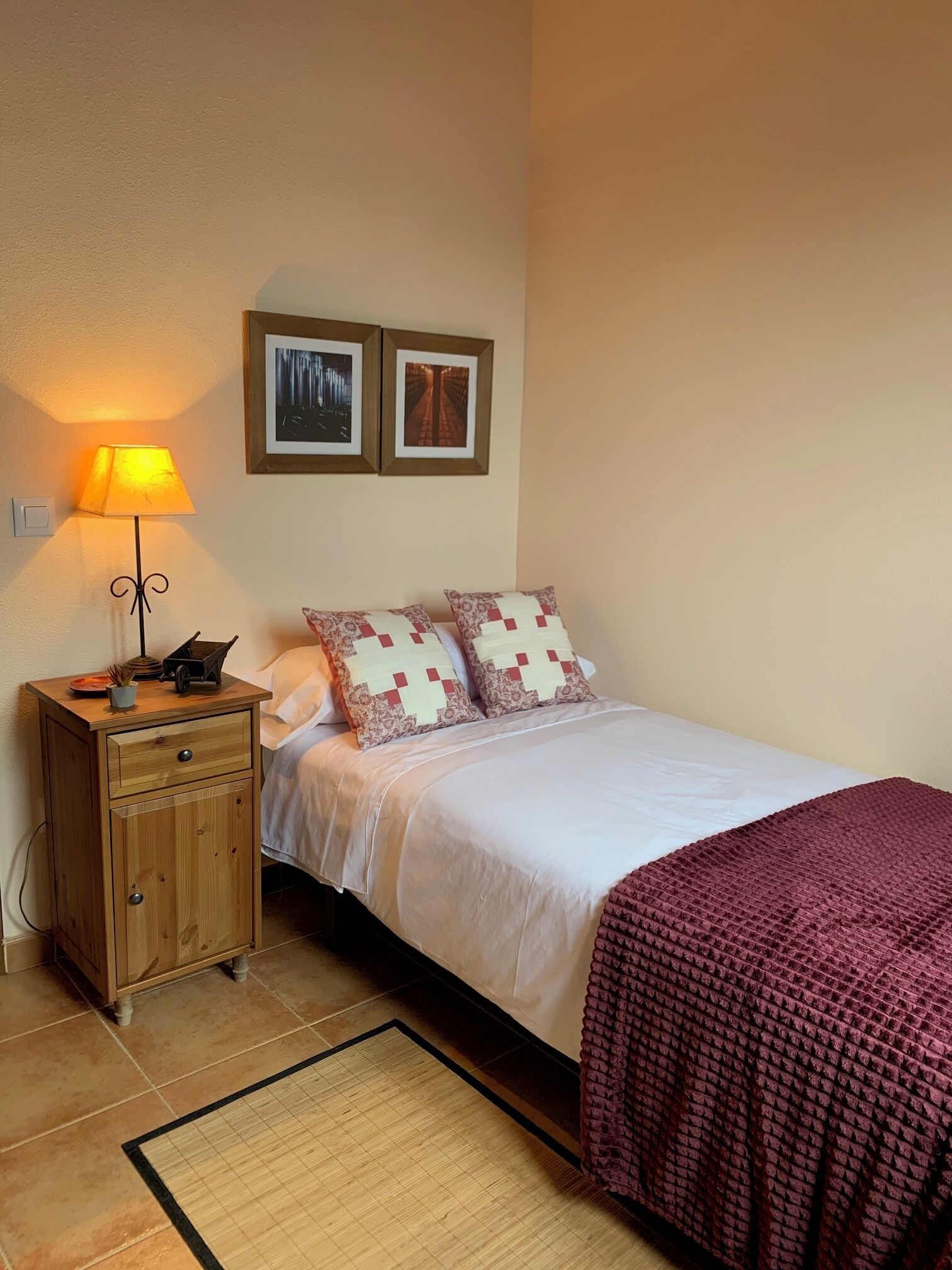 A neatly arranged bedroom with a bed, a bedside table with a lamp, a small cabinet, and two framed pictures on the wall.