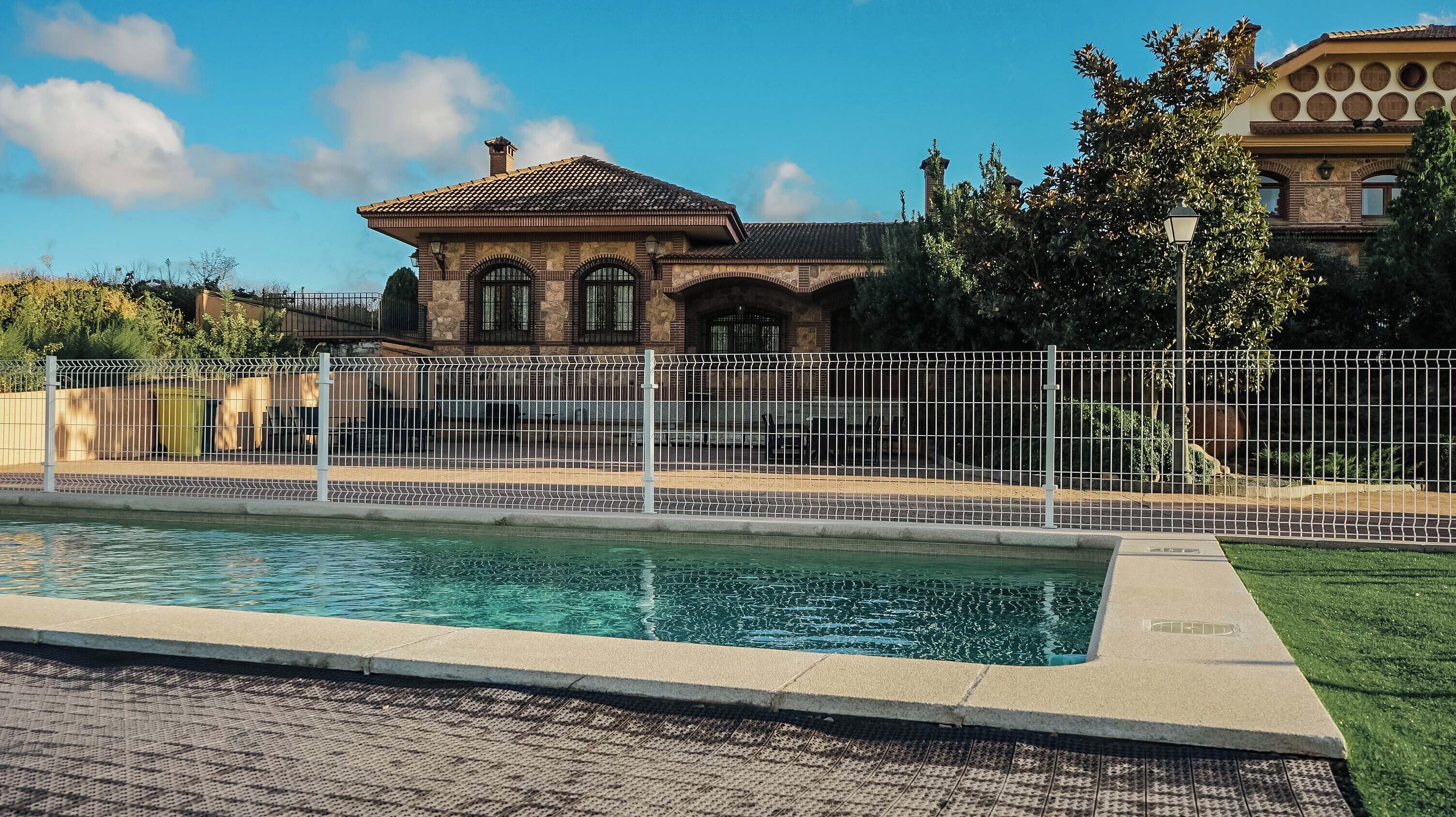 Una piscina con una cerca blanca, un edificio de piedra con ventanas de arco y un cielo azul despejado.