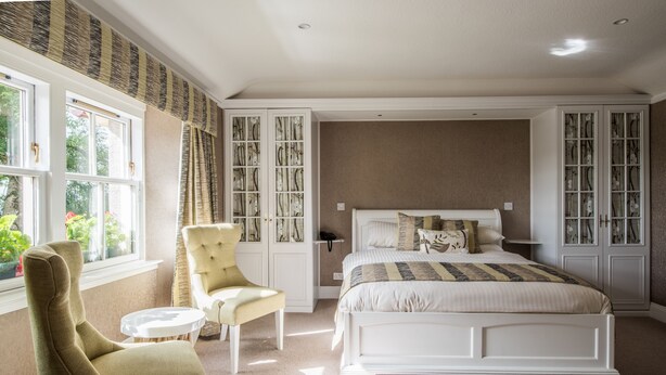 A bedroom with a large bed, a chair, a small table, and a window with curtains. - The Redgarth (Inverurie)