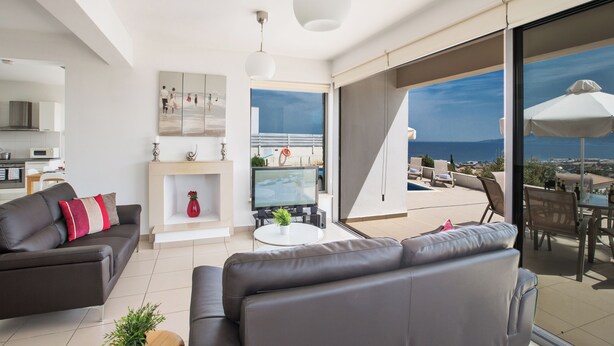 A modern living room with a sofa, a TV, and a dining area with a view of the sea. - Villa Abbie, Panoramic Sea Views (Polis)