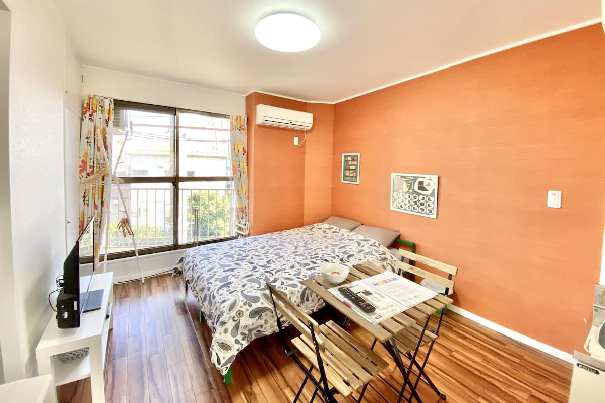 Una habitación pequeña y luminosa con una cama, una mesa de madera y un ventanal con cortinas.