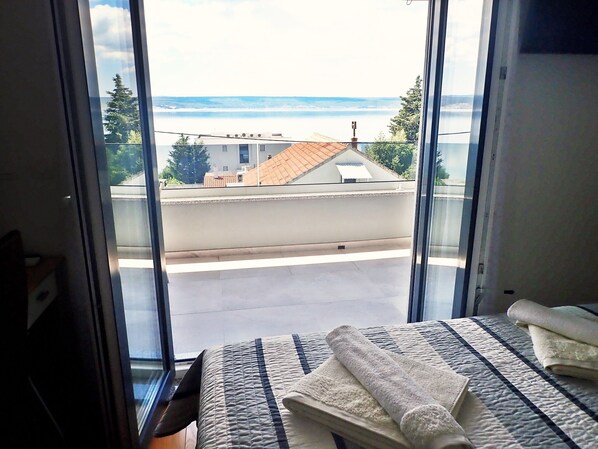 A bedroom with a bed, a window with a view of the sea, and a balcony with a railing.