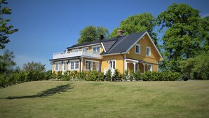Een gele, twee verdiepingen tellende woning met een balkon, omringd door een gazon en bomen.