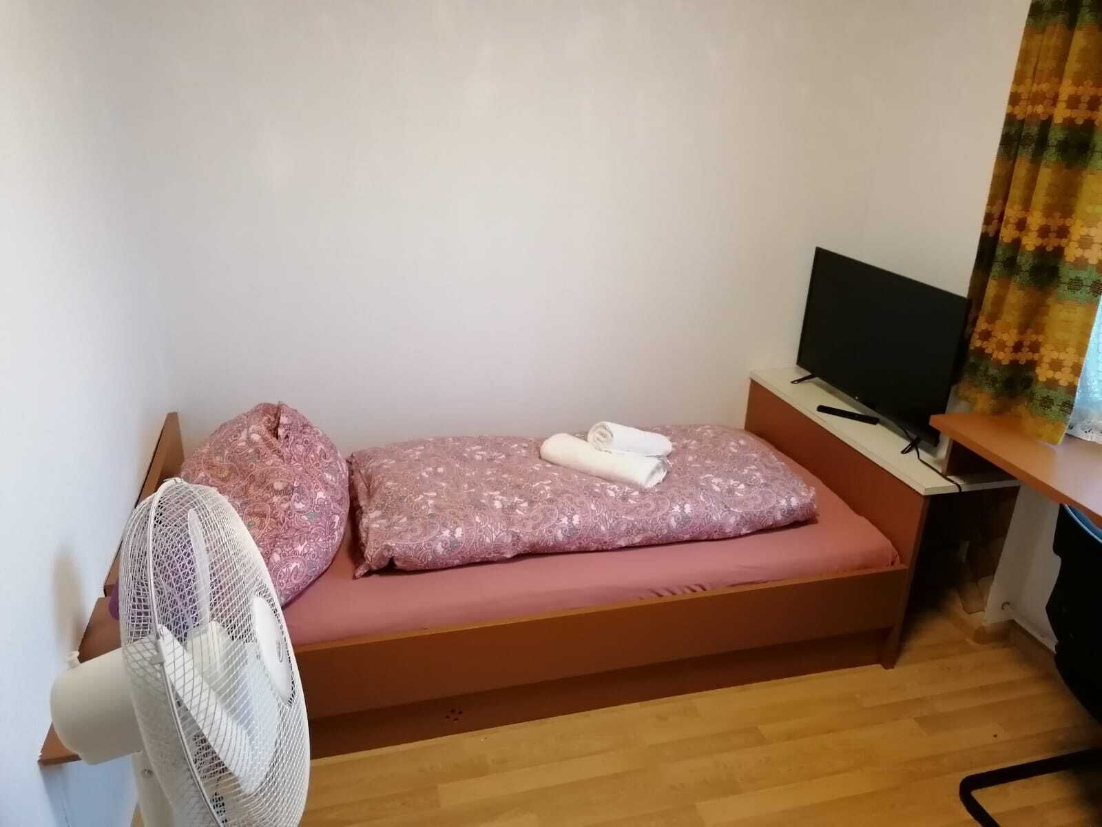 Una habitación pequeña con cama individual, piso de madera, cama con colchón y almohada rosa, ventilador blanco, televisor sobre una mesa de madera y cortina con estampado floral.