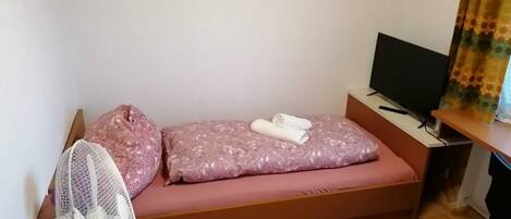 Una habitación pequeña con cama individual, piso de madera, cama con colchón y almohada rosa, ventilador blanco, televisor sobre una mesa de madera y cortina con estampado floral.