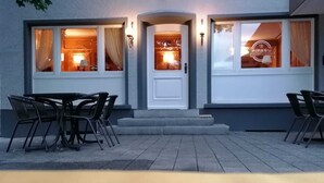 A building with a white door, two windows with curtains, and a sign reading "PIPABU Pizza Pasta Burger." There are four chairs in front of the building. - PiPaBu Restaurant & Hotel (Rheinfelden (Baden))