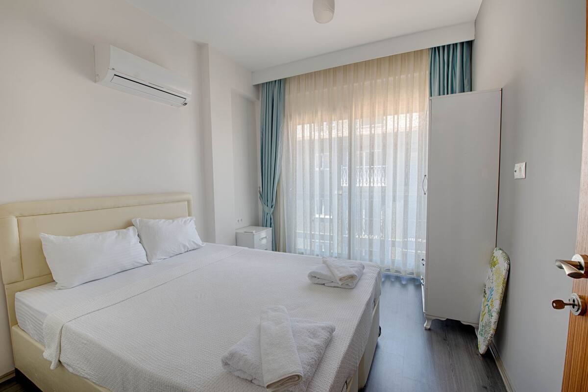 A hotel room with a large bed, a bedside table, an air conditioning unit, a wardrobe, and a window with curtains.