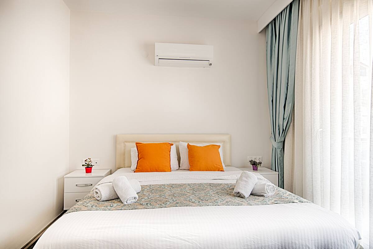 A neatly made bed with white and orange bedding, a small bedside table with a red pot, and a white air conditioning unit on the wall.