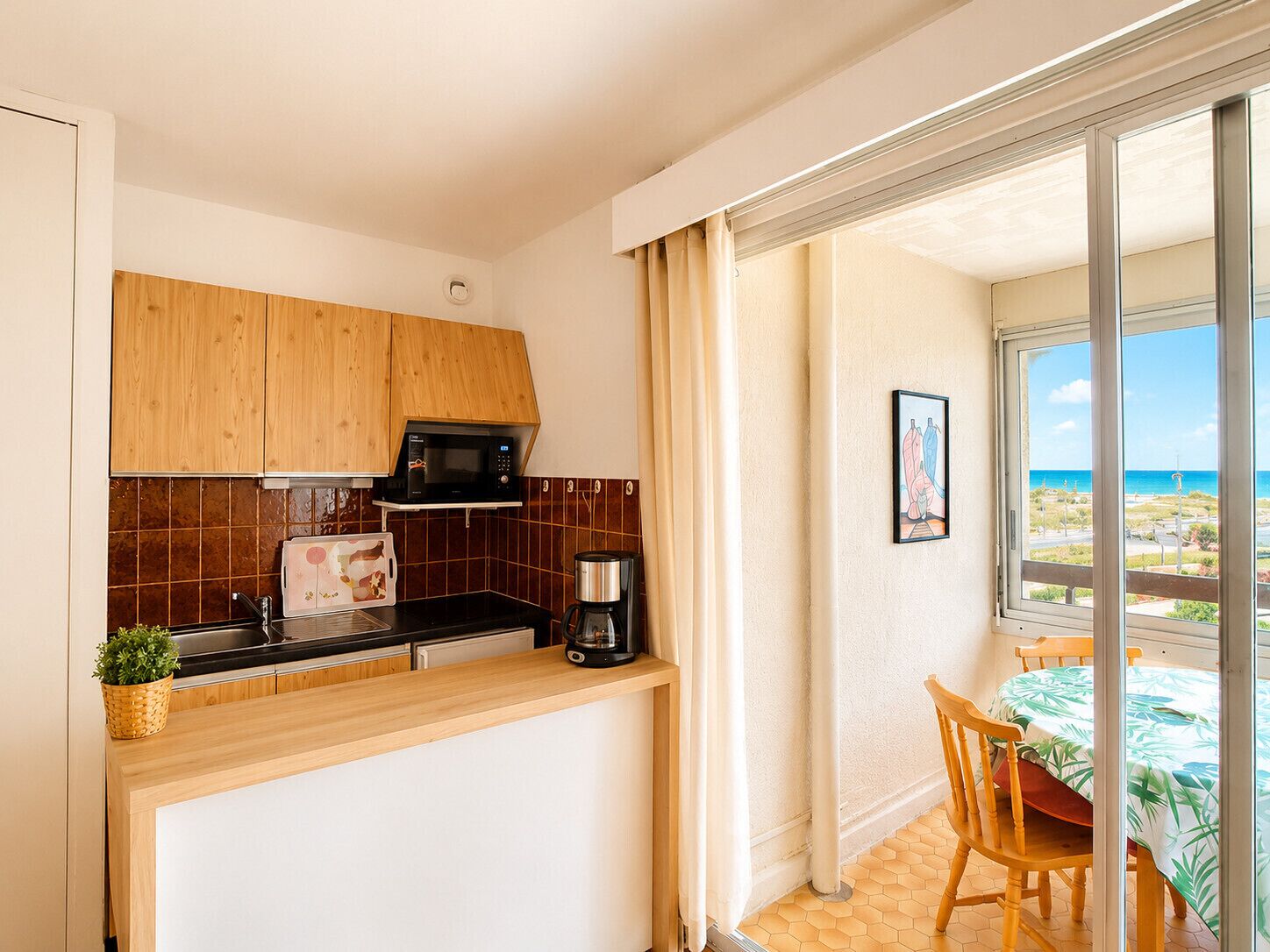 A compact kitchen with wooden cabinets, a microwave, a coffee maker, and a view of the sea through a glass door.
