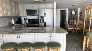 A kitchen with white cabinets, a stainless steel refrigerator, a microwave, and a coffee maker. There are three wicker stools with green cushions. - Direct Oceanfront Condo remodeled with Breathtaking Views from two private decks (Ocean City)