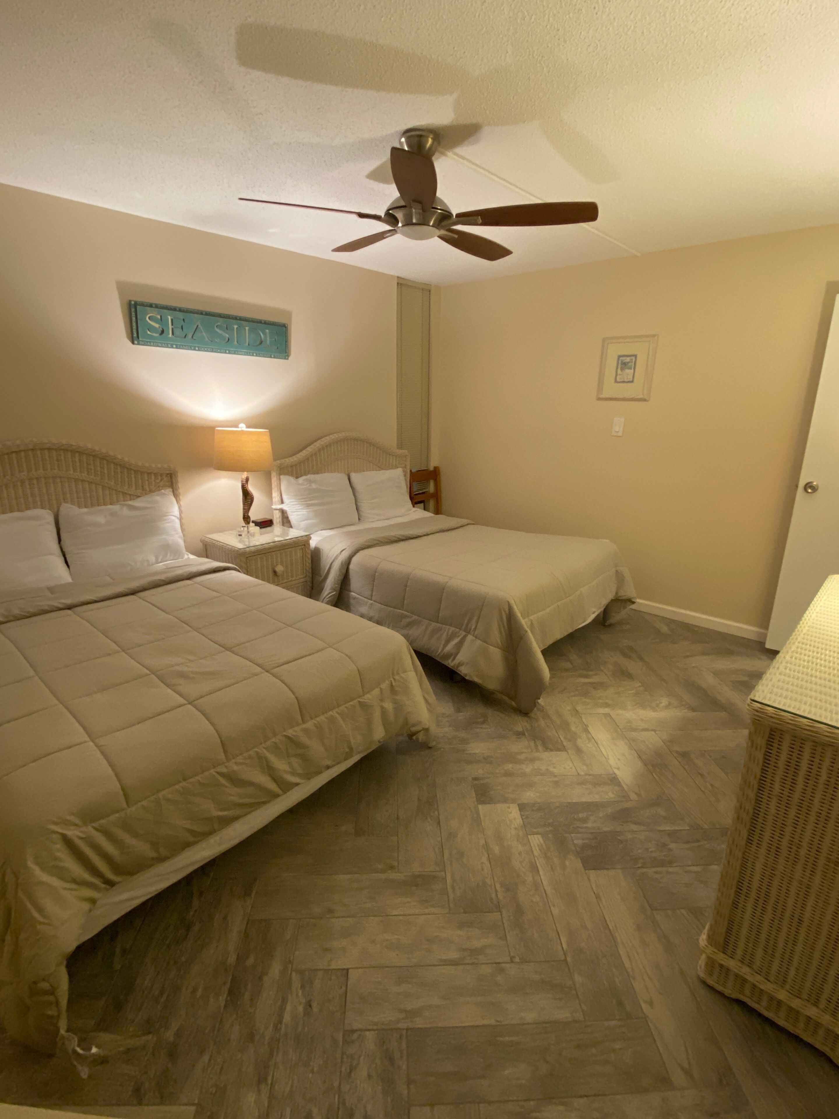 Ein Hotelzimmer mit zwei Betten, einem Deckenventilator, einem Nachttisch mit Lampe und einem Korbstuhl.