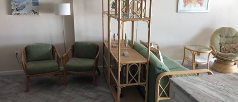 Una habitación con una cama de ratán, dos sillas con cojines verdes, una mesita con lámpara y obras de arte en las paredes.