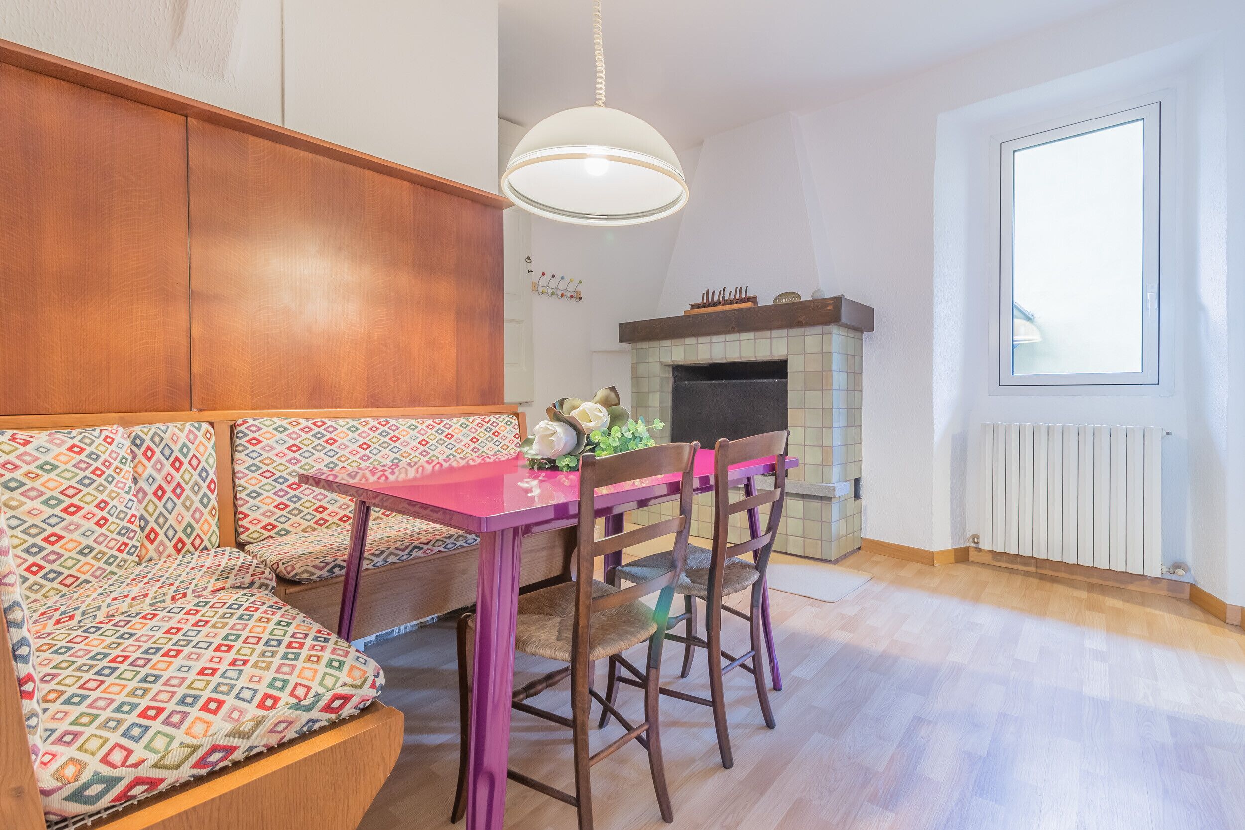 A cozy room with a wooden bench, a pink table, and two chairs. There's a fireplace with a tiled surround and a window with blinds.