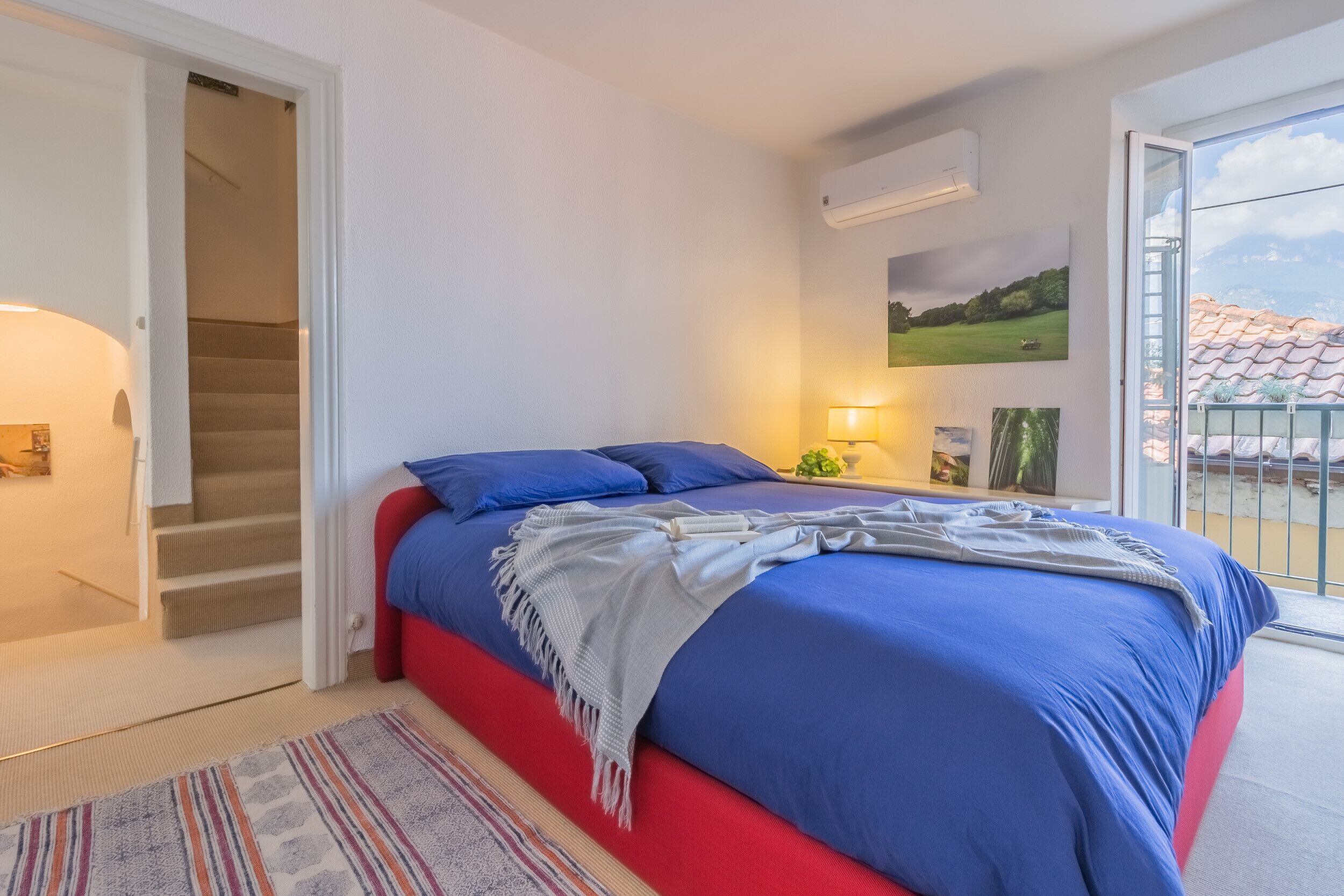 Un dormitorio con cama, mesita de noche, lámpara, un cuadro y vistas al exterior.