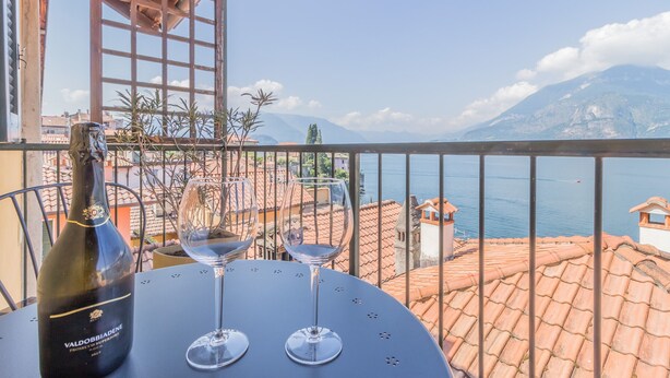 Ein Tisch mit einer Weinflasche und zwei Gläsern auf einem Balkon mit Blick auf eine Berglandschaft und ein Gewässer.