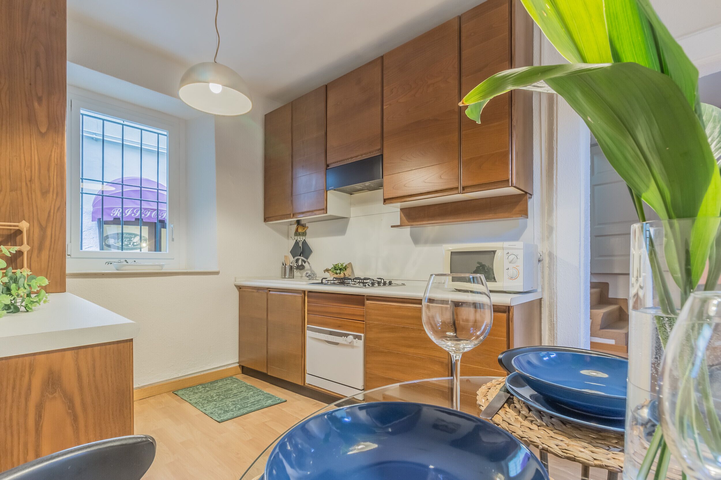 A modern kitchen with wooden cabinets, a microwave, and a dining area with a glass table and blue plates.