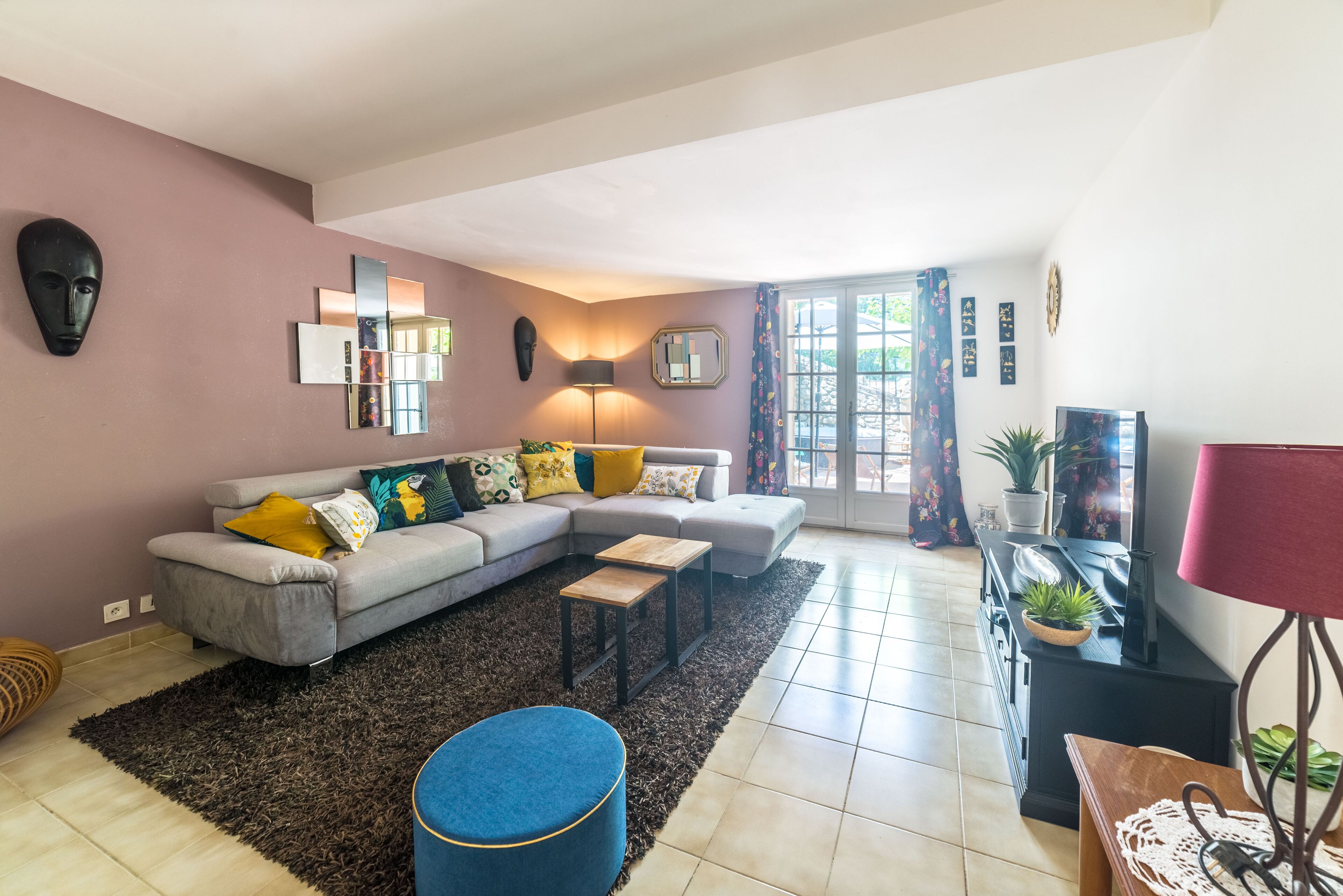 Ein Wohnzimmer mit einem grauen Ecksofa, einem blauen Hocker, einem Couchtisch aus Holz und einem Fernseher auf einem schwarzen Standfuß.