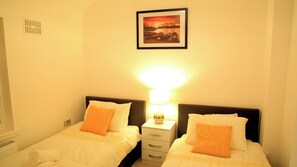 A hotel room with two beds, a nightstand with a lamp, a picture on the wall, and a window. - A Modern, Comfy Newly Remodeled 2bd House (Dagenham)