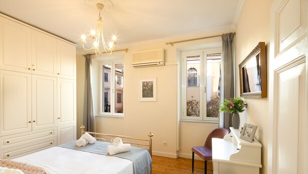 A bedroom with a bed, a desk, and a chair. There is a window with curtains, a chandelier, and an air conditioning unit on the wall. - San Nicolo dei Vecchi Casa  by CorfuEscapes (Corfu)