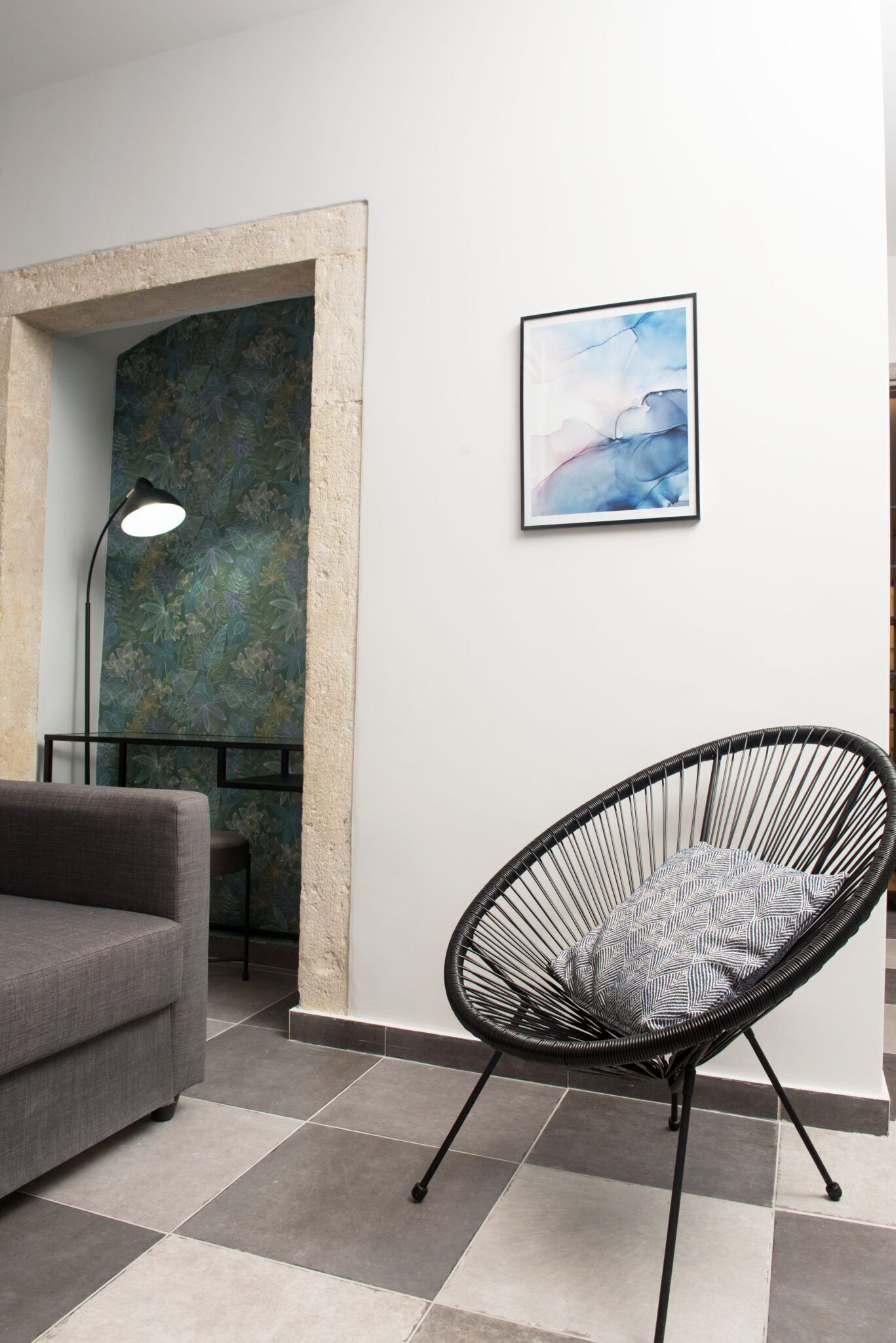 A modern living room with a grey sofa, a black wireframe chair, a floor lamp, and a framed abstract painting on the wall.