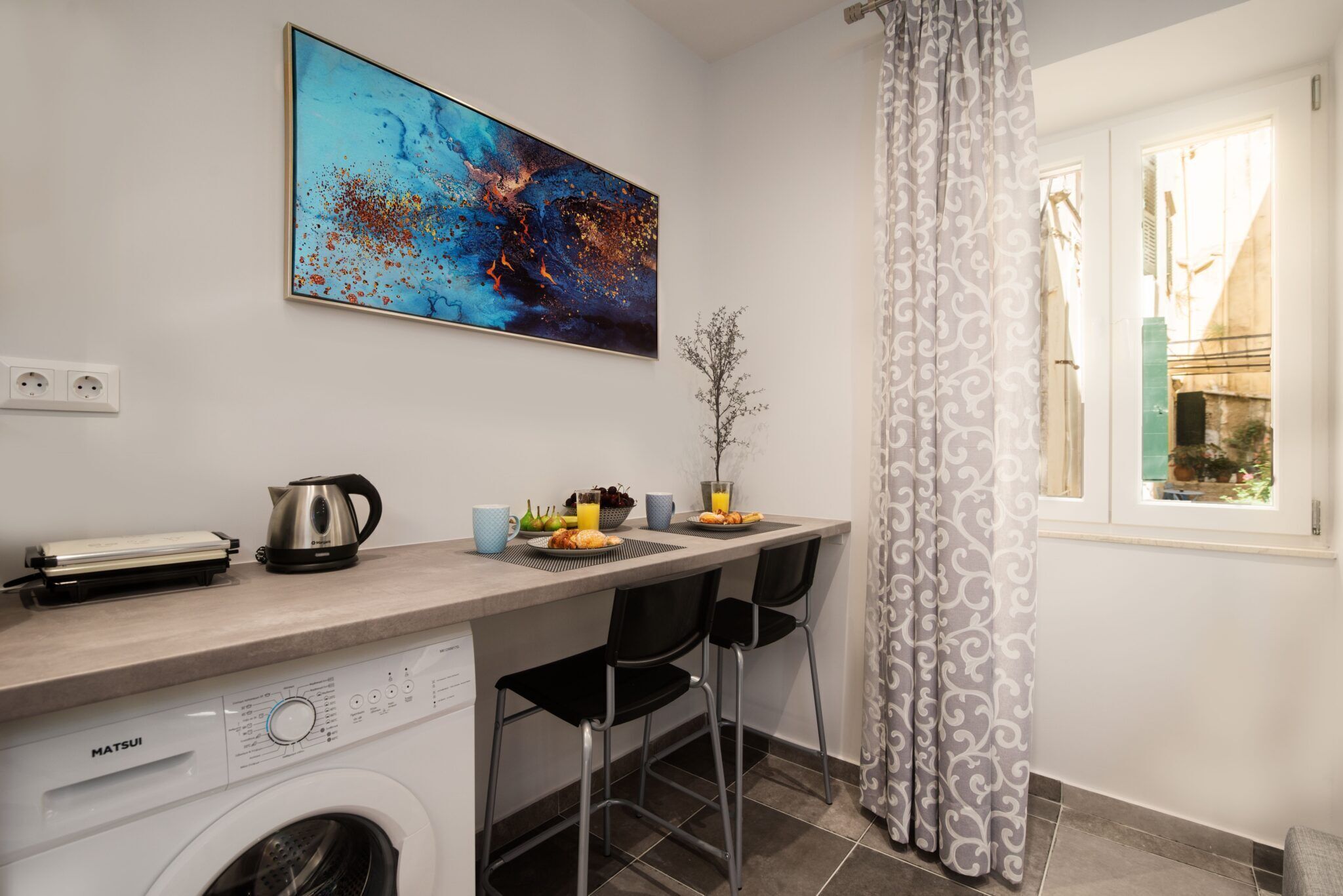 Une cuisine compacte équipée d’un lave-linge, d’une machine à café et d’un grille-pain. Il y a une fenêtre avec des rideaux et un tableau au mur.