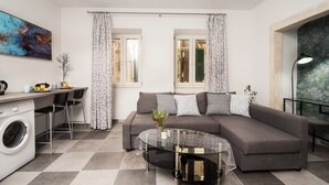 A modern living room with a grey sofa, a glass coffee table, and a washing machine. - Eden Apartments  by CorfuEscapes (Corfu)