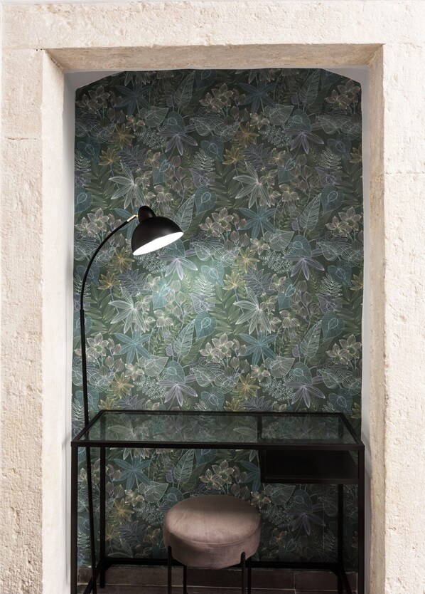 A corner with a green floral wallpaper, a black desk with a chair, and a black floor lamp.