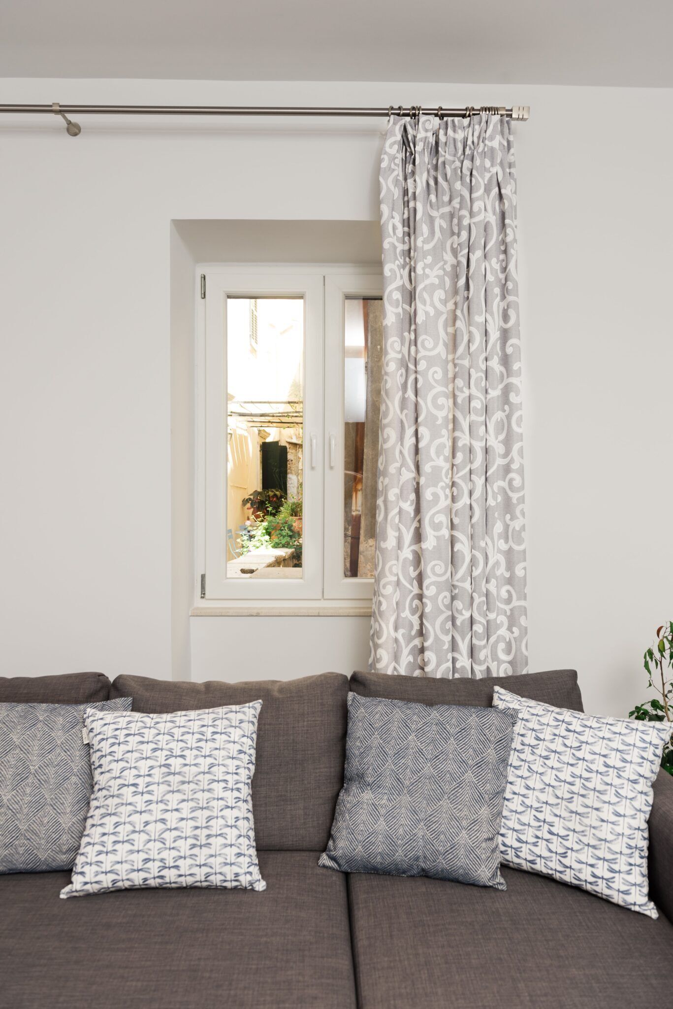 A living room with a grey sofa, patterned cushions, a window with a view of a garden, and a curtain with a white and grey pattern.