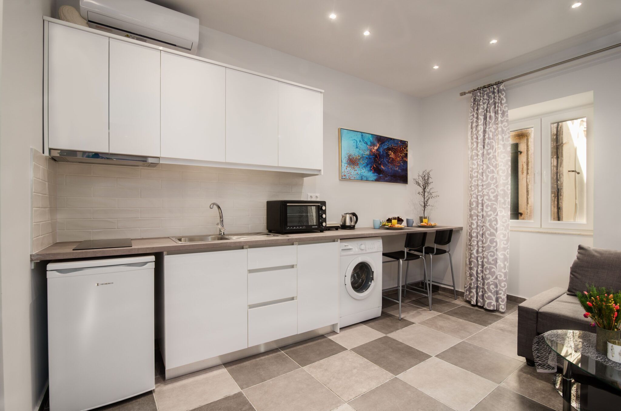 Cocina compacta con armarios blancos, lavavajillas, lavadora, microondas y cafetera. Cuenta con un comedor con mesa y sillas, y una ventana con cortinas.