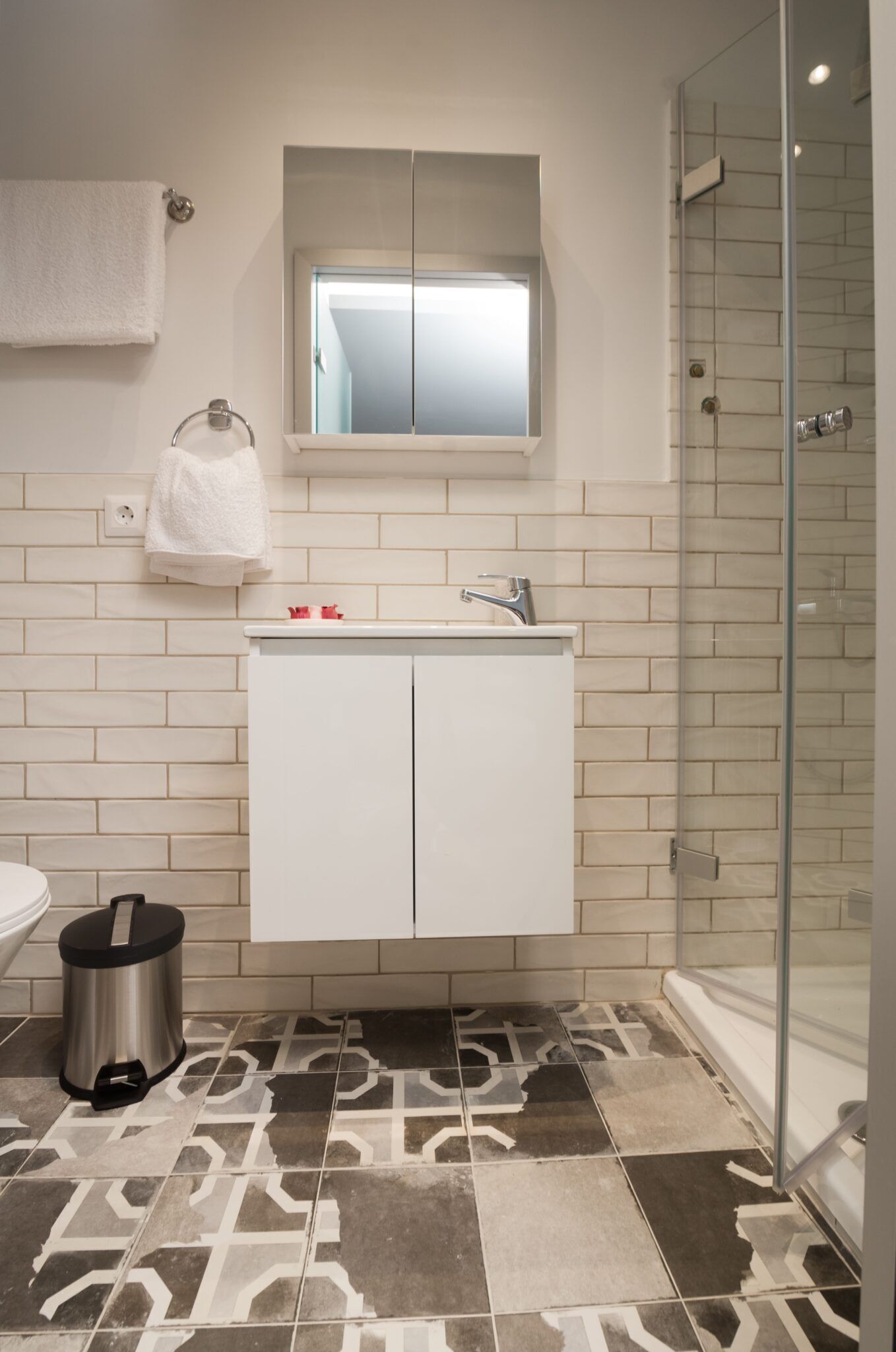 Un baño con pared de azulejos blancos, un ventanal sobre el lavamanos, cabina de ducha con mampara de vidrio y suelo estampado.