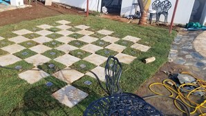 A paved outdoor area with a checkered pattern of grass and stone tiles, surrounded by a low wall and a few chairs.