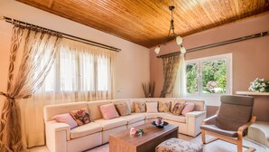 Un salon avec un canapé d’angle beige, une table basse en bois, un pouf à motifs, un fauteuil marron et une grande fenêtre donnant sur un paysage verdoyant.