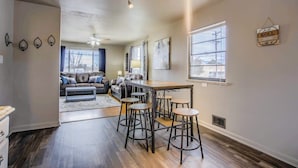 A modern kitchen with a dining area, featuring a bar stool set, a large window, and a ceiling fan.