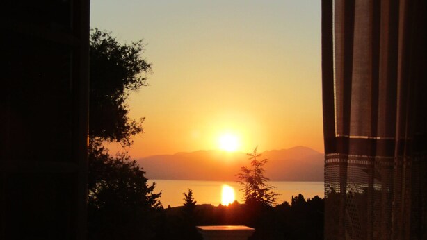 A balcony with a view of a sunset over a body of water, framed by trees and mountains. - Villa Southern Comfort by CorfuEscapes (Corfu)