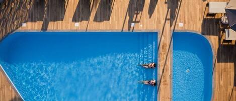 A rectangular swimming pool with two people swimming, surrounded by wooden decking and outdoor furniture.