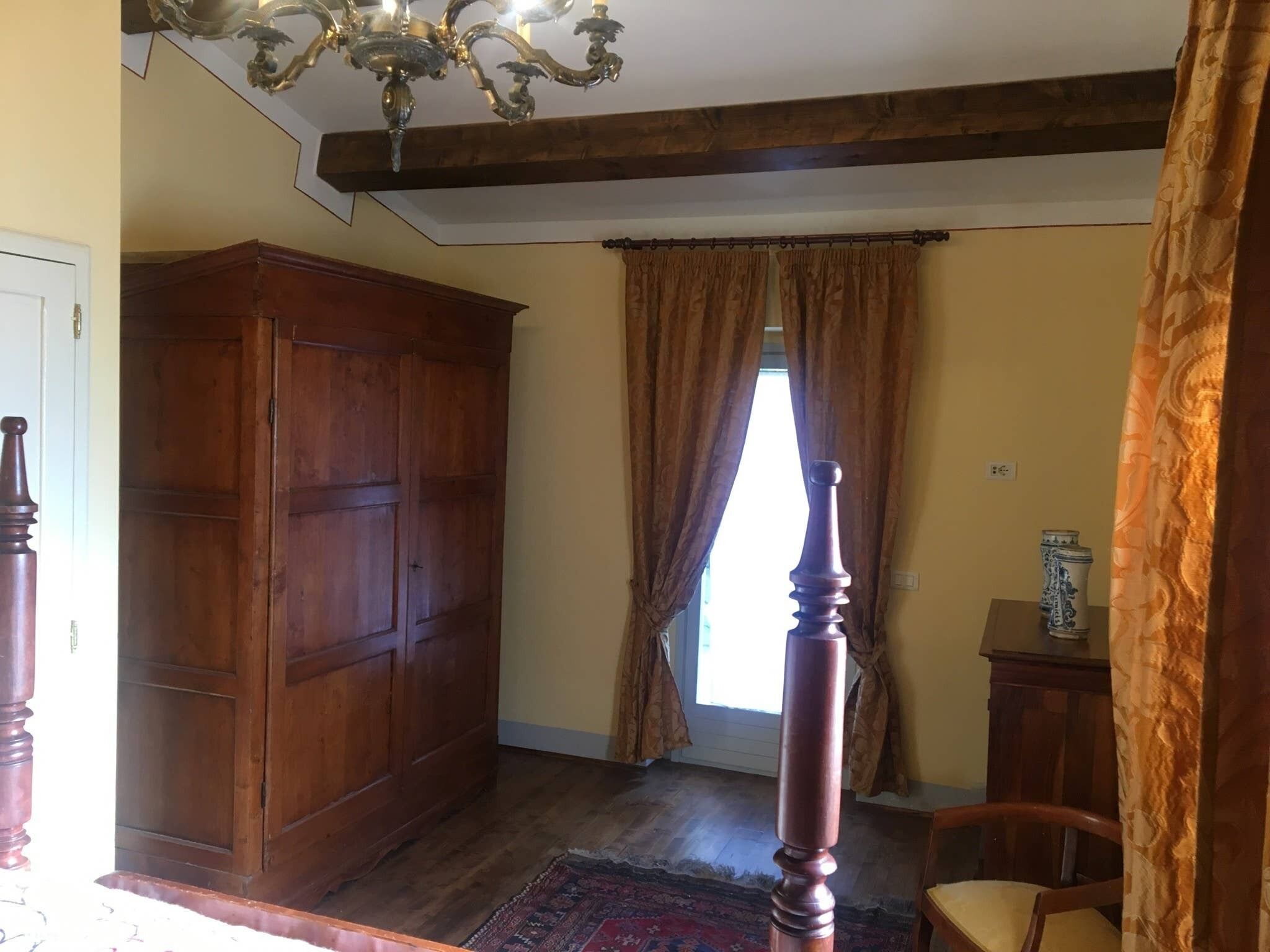 A room with a large wooden wardrobe, a wooden chair, a small table with a vase, and a window with curtains.