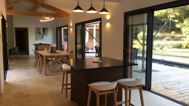Een moderne keuken met een centraal eiland, barkrukken, hanglampen en grote ramen met uitzicht op de omgeving.