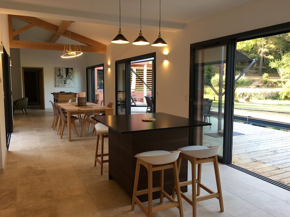 Une cuisine moderne dotée d’un îlot central, de tabourets de bar, de luminaires suspendus et de grandes fenêtres offrant une vue sur l’extérieur.